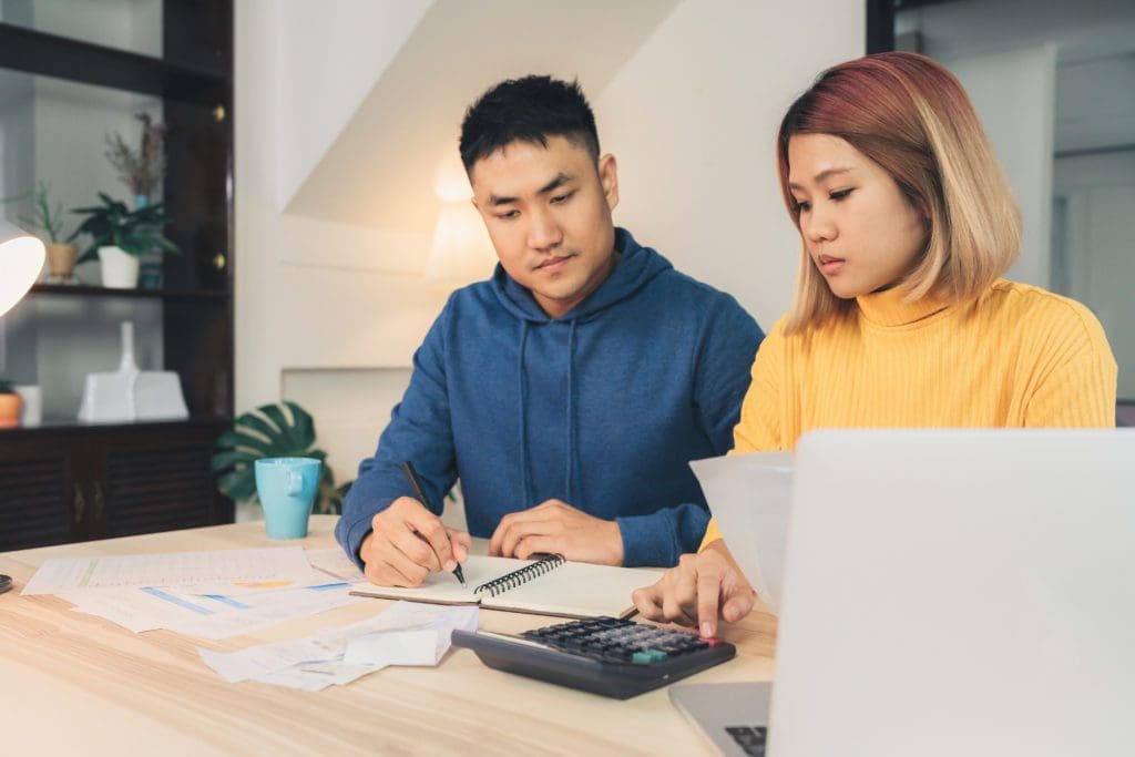 Young couple managing finances, reviewing their bank accounts using laptop computer and calculator at modern home.