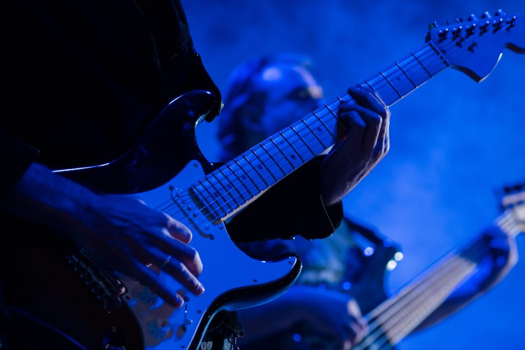 A live guitarist's hands are shown riffing to a rock and roll song under blue light.